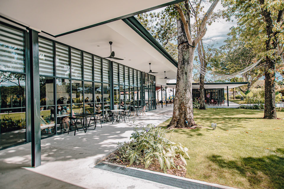 Modern outdoor cafe with glass walls, seating on the patio, surrounded by trees and greenery on a sunny day.