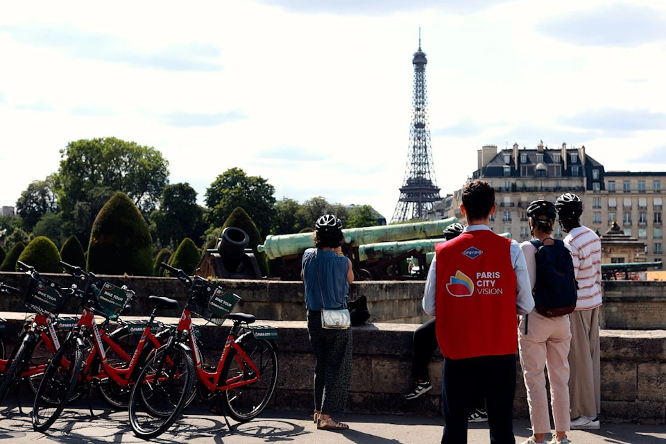 Tourists with bicycles standing near a historic cannon, facing the Eiffel Tower in Paris. A person wears a "Paris City Vision" vest.