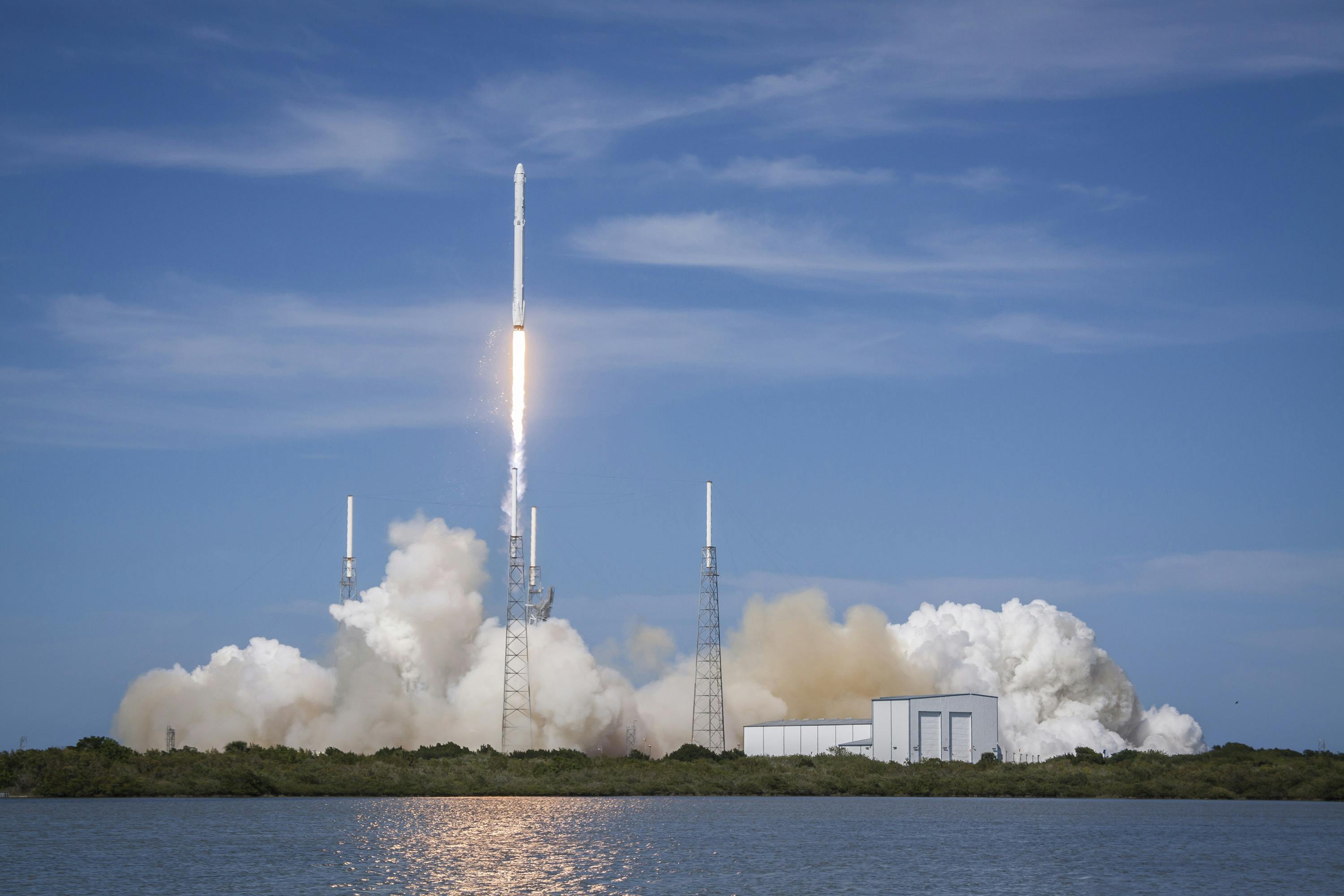 A rocket launches into a blue sky, emitting smoke and fire, surrounded by support structures and a building near a body of water.
