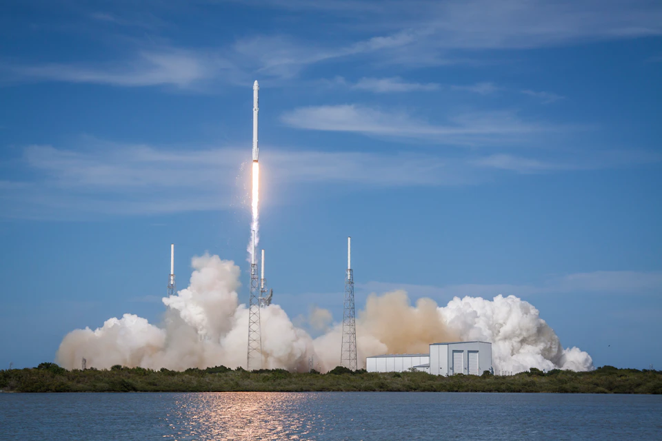 A rocket launches into a blue sky, emitting smoke and fire, surrounded by support structures and a building near a body of water.