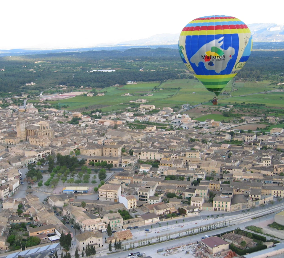 Colorful hot air balloon with the word "Mallorca" floats above a town with clustered buildings, open fields, and distant mountains.