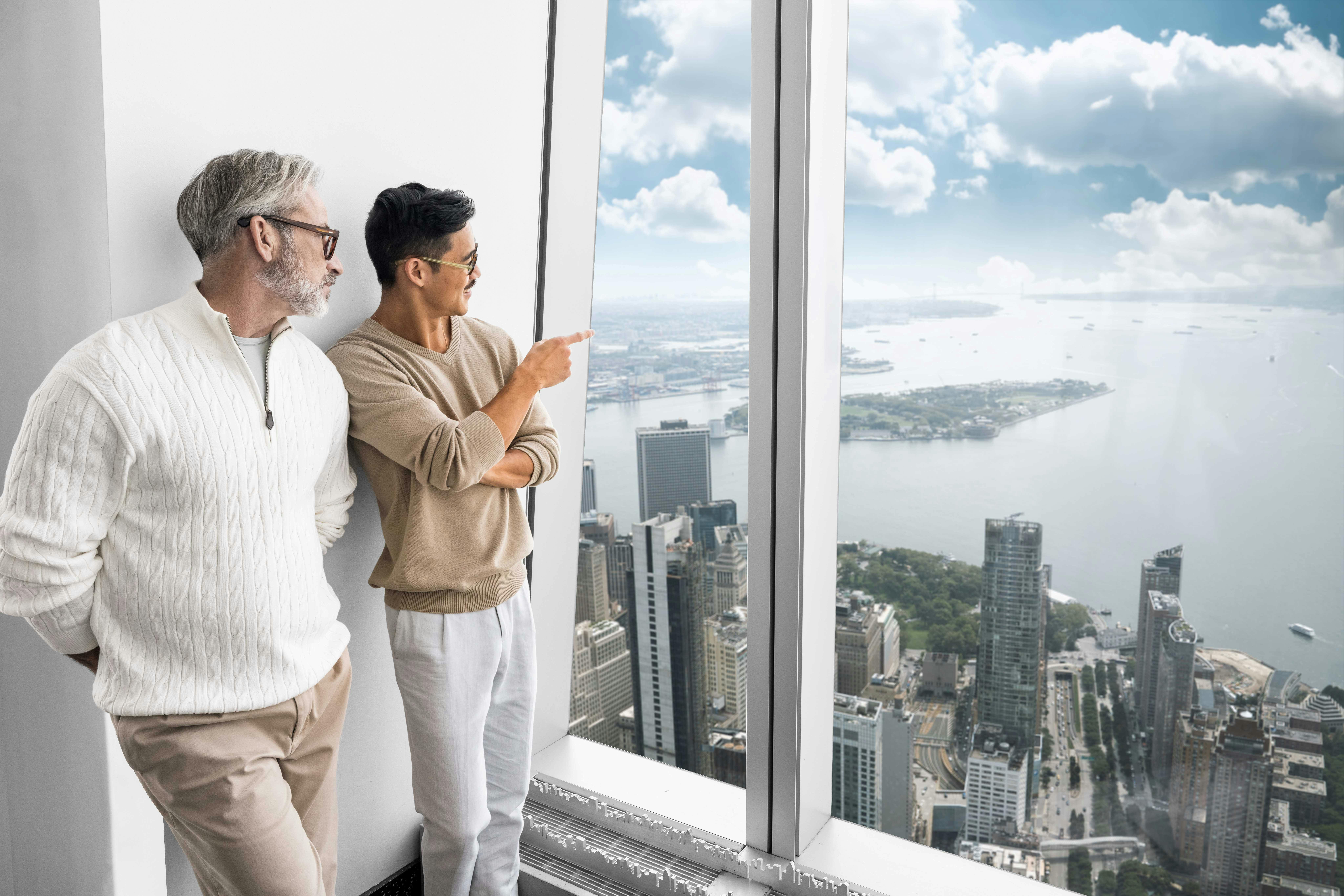 Two men stand indoors by a large window overlooking a cityscape with tall buildings, water, and a partly cloudy sky.