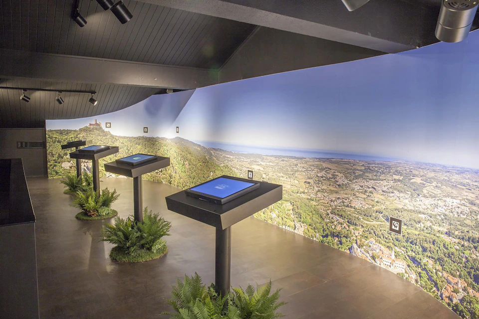An exhibit room featuring digital screens on stands with a panoramic landscape mural on the wall and decorative plants on the floor.
