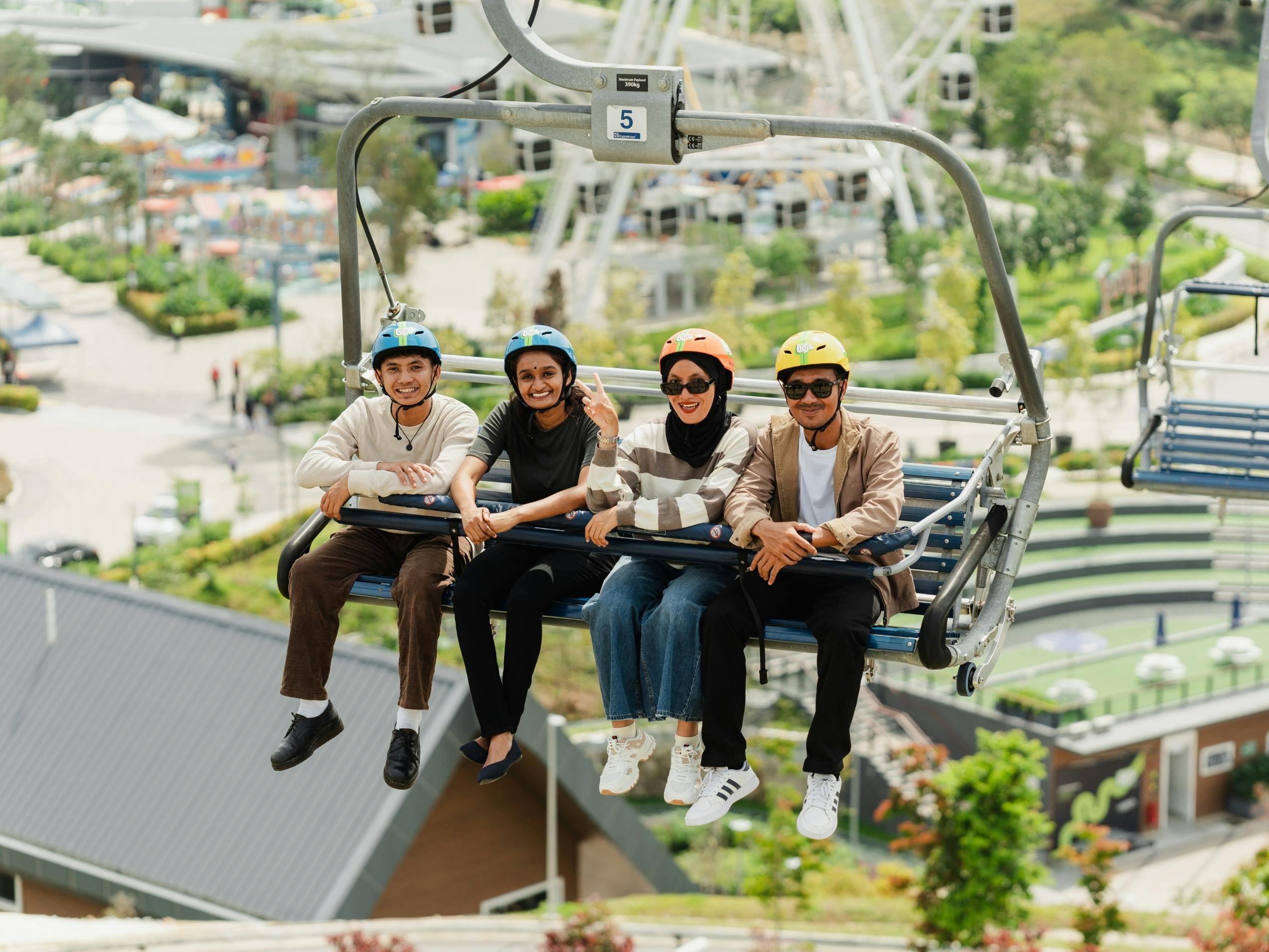 Four people sitting on a chairlift, smiling and wearing helmets, with a park and buildings visible in the background.