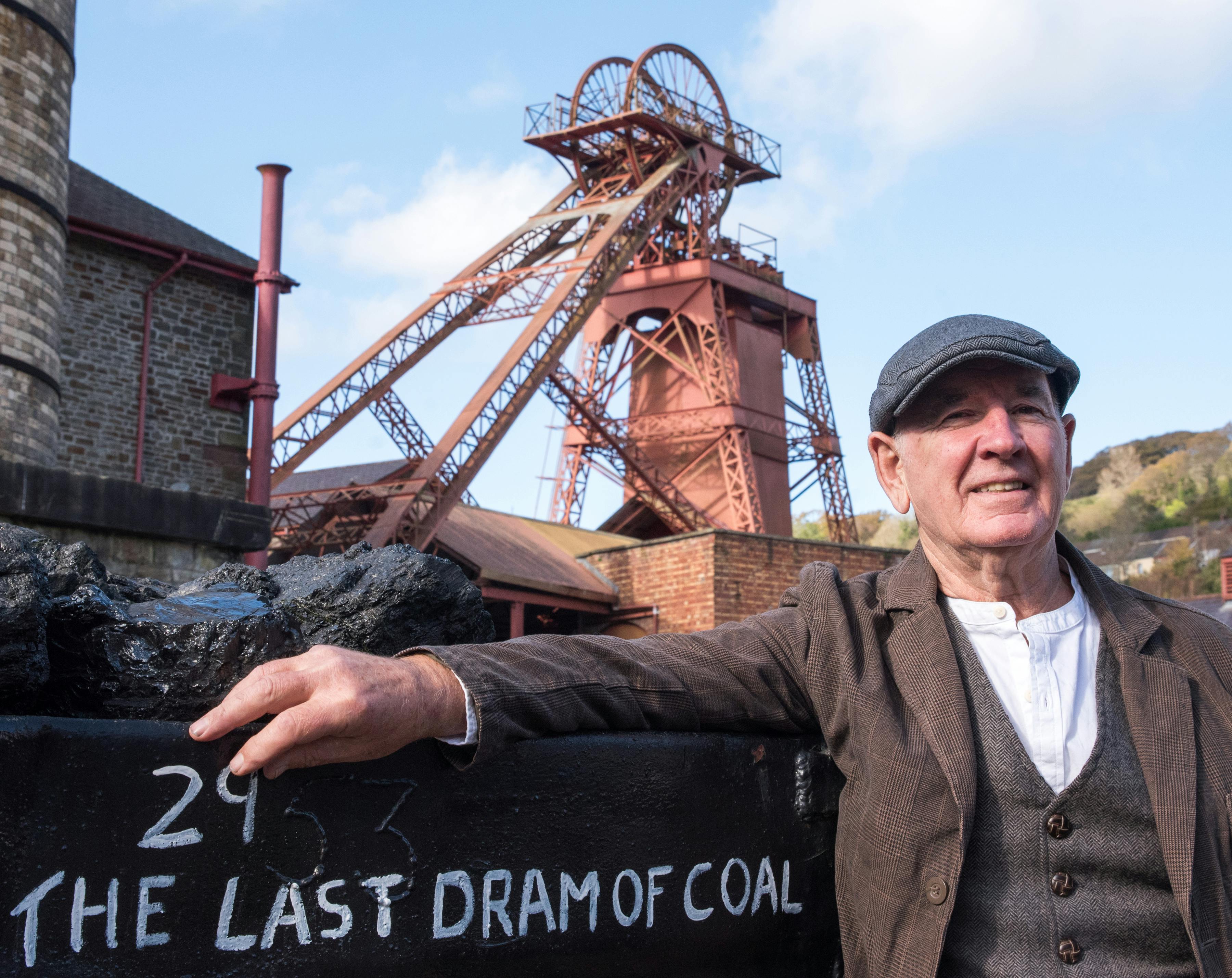 빨간색 광산 구조물을 배경으로 "석탄의 최후의 잔" 이라고 적힌 용기 옆에 모자를 쓴 남성이 서 있습니다.