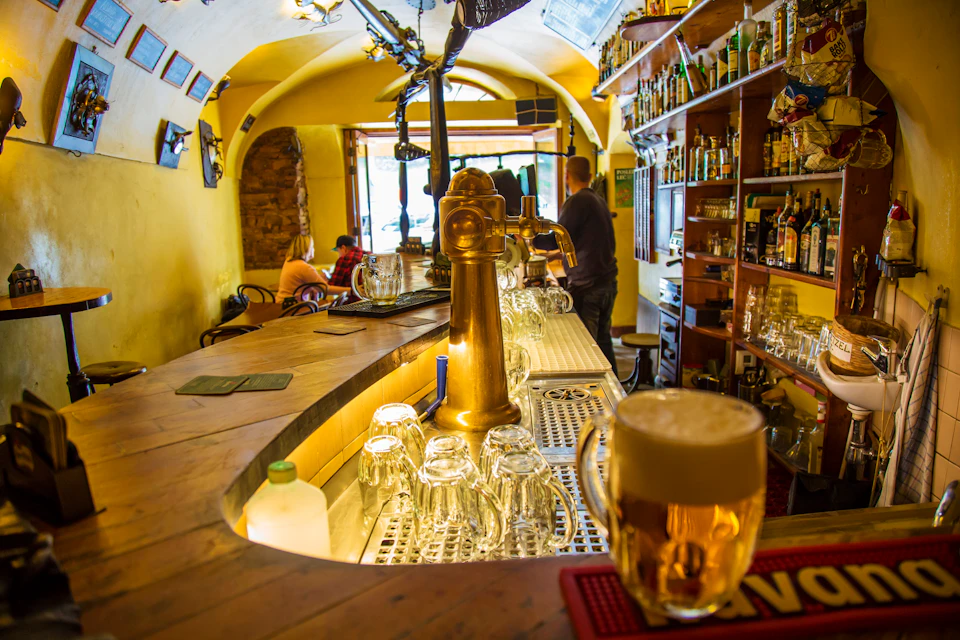 Cozy pub interior with a curved wooden bar, beer taps, empty glasses, and a poured beer. Patrons sit and chat near the entrance.