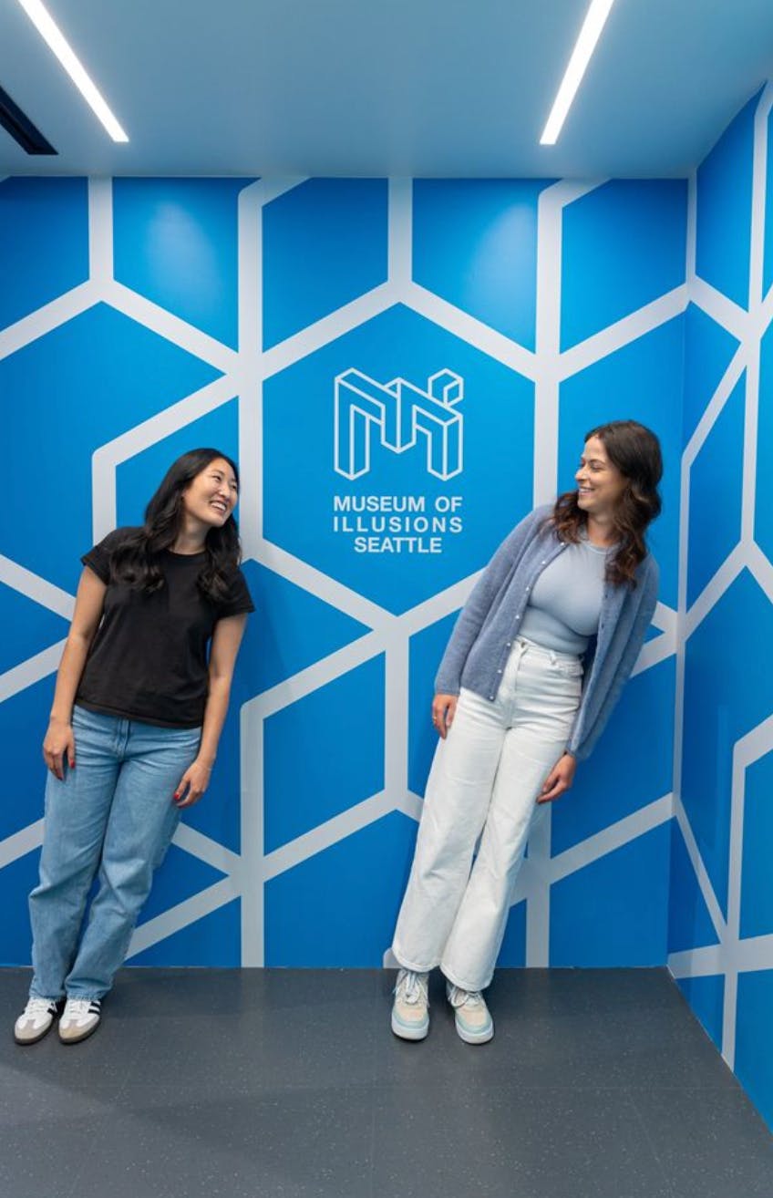Zwei Personen stehen vor einer blauen Wand mit geometrischen Mustern und der Aufschrift "Museum of Illusions Seattle" und lächeln sich an.