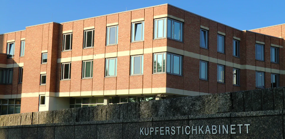 Brick building with multiple windows above a concrete structure that has "KUPFERSTICHKA" partially visible. Clear blue sky.