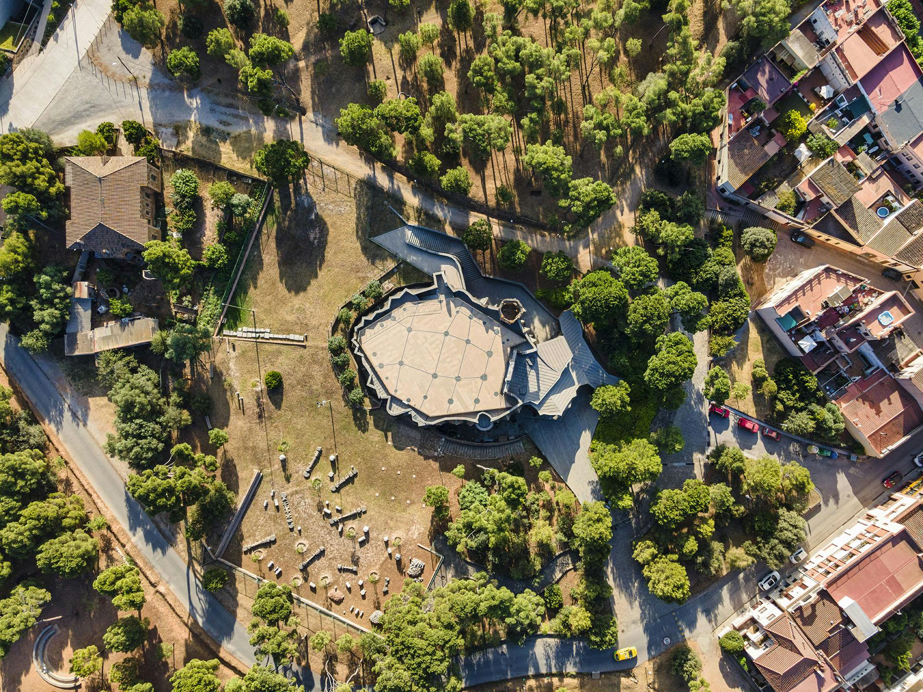Aerial view of a hexagonal building surrounded by trees, pathways, and a nearby residential area with parked cars.