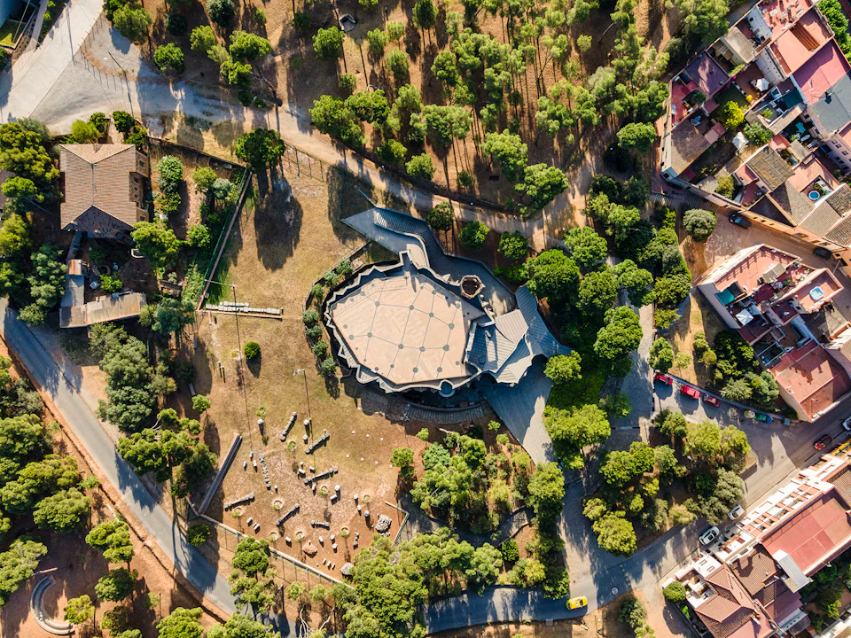 Aerial view of a hexagonal building surrounded by trees, pathways, and a nearby residential area with parked cars.