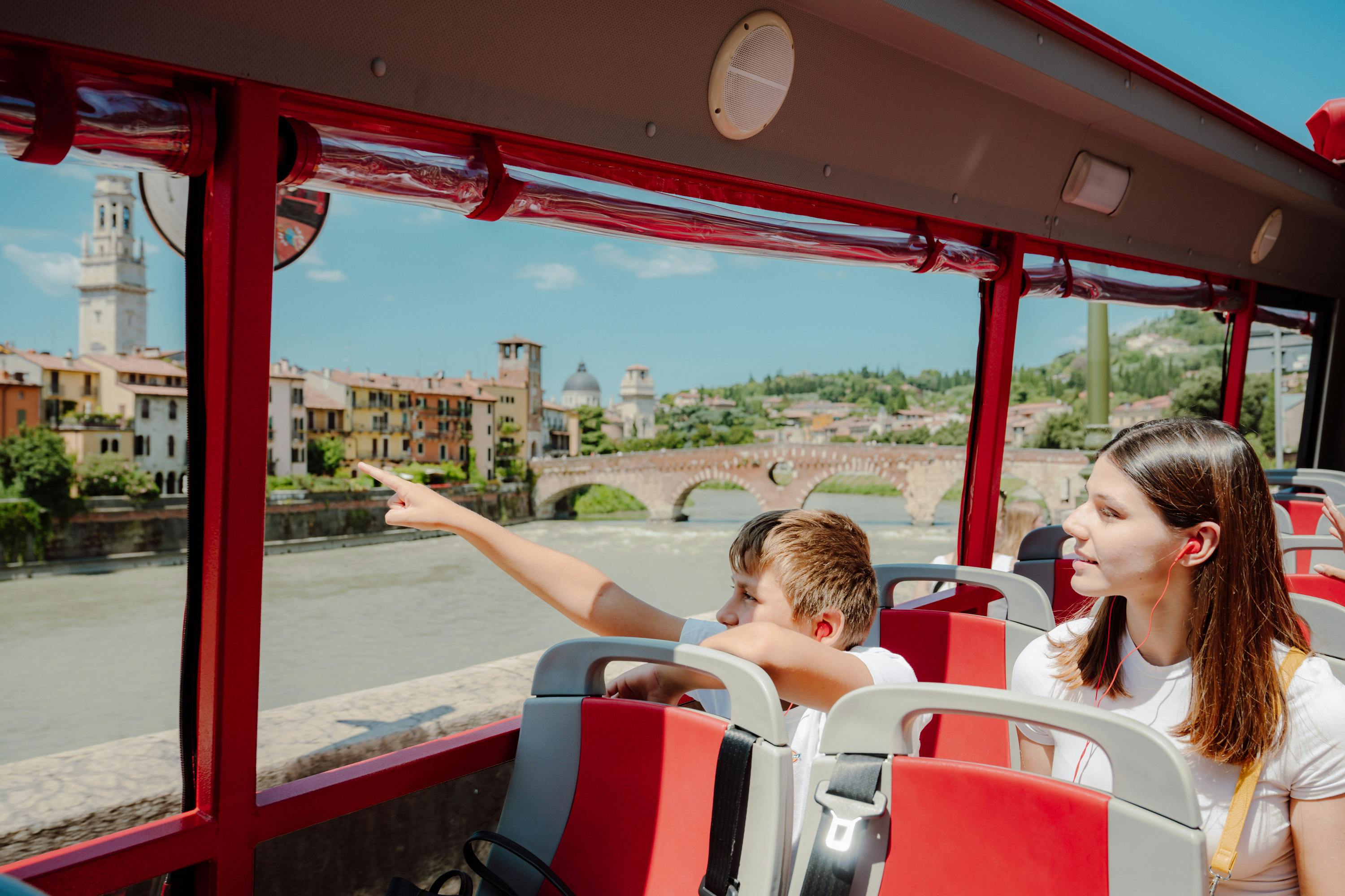 Due persone su un autobus turistico rosso, una delle quali indica una vista panoramica su un fiume e un ponte storico in pietra.