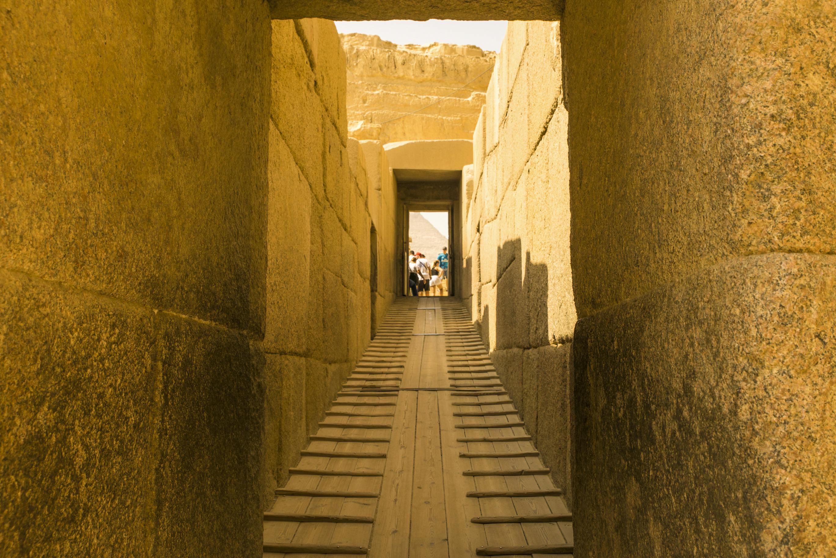 A stone corridor with a wooden ramp leads to a group of people in the distance, with ancient ruins visible in the background.