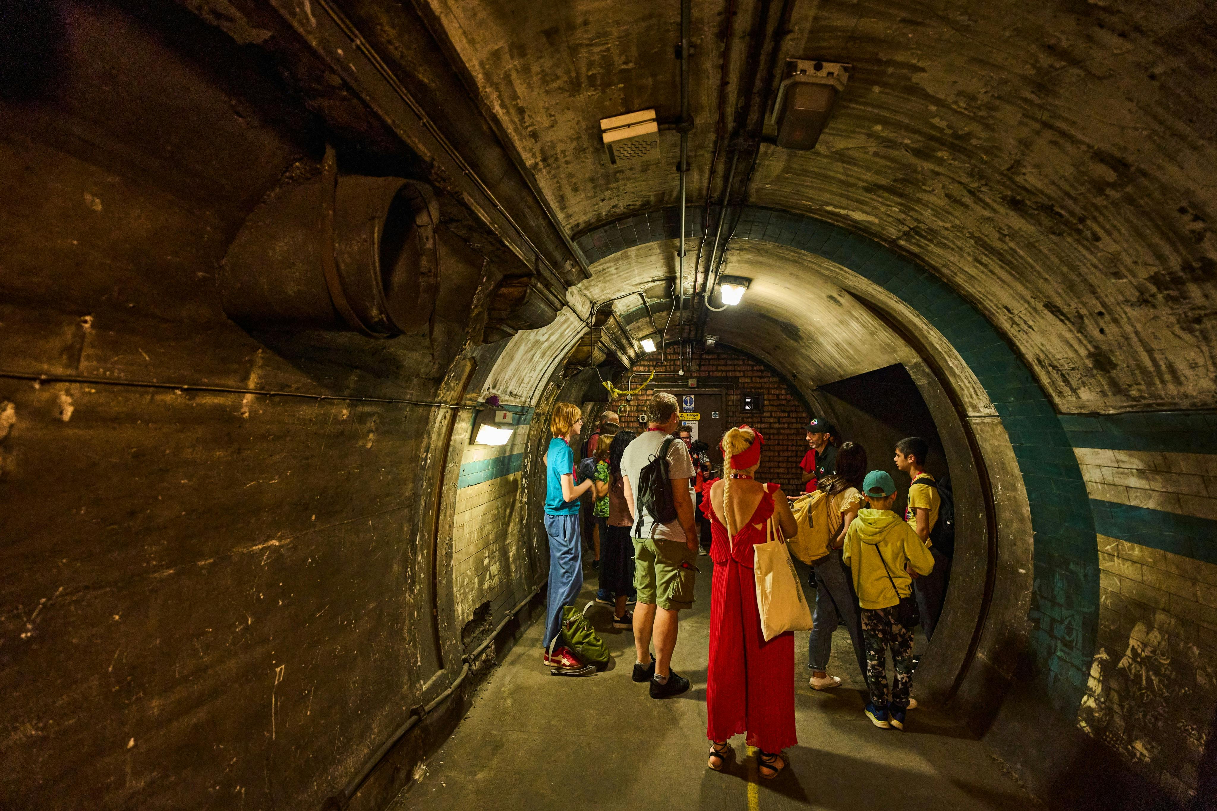 A group of people gather in an underground tunnel with curved walls, dim lighting, and visible utility features.