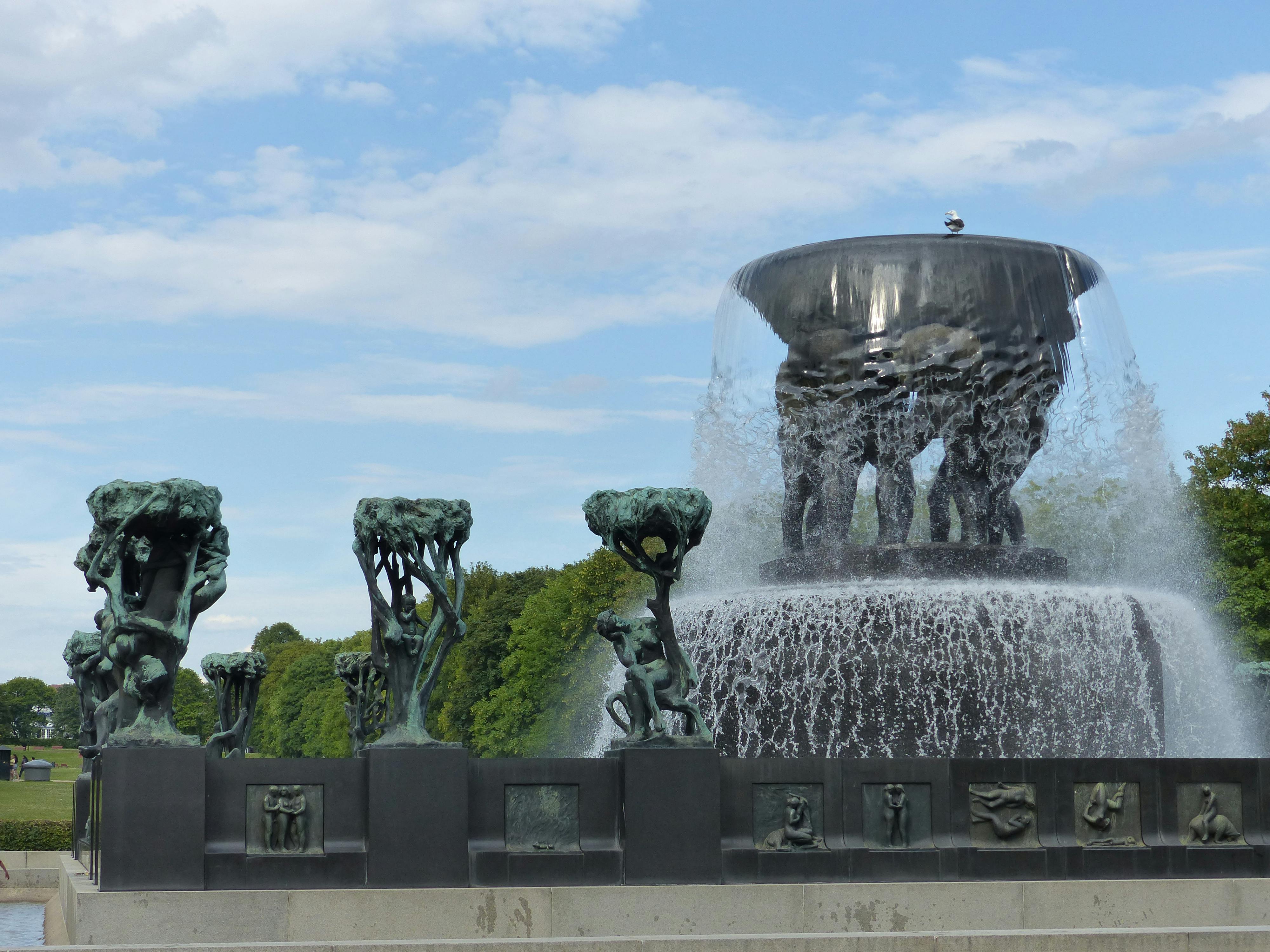 Χάλκινα γλυπτά από φιγούρες και δέντρα ευθυγραμμίζουν μια μεγάλη βρύση νερού σε ένα πάρκο με γαλάζιο ουρανό και σύννεφα στο παρασκήνιο.