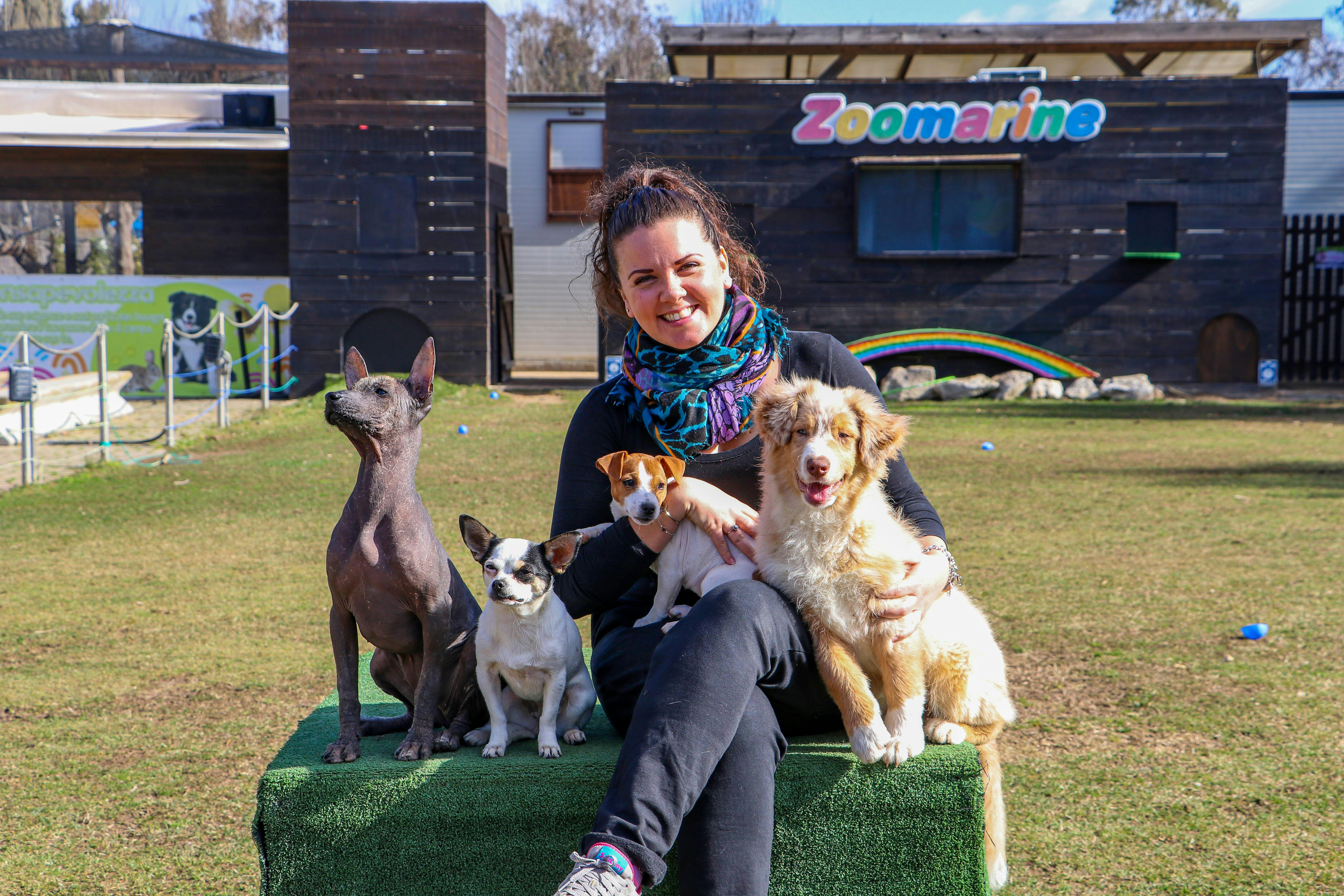 Žena sedí na zelené plošině, usmívá se, obklopena čtyřmi psy různých plemen před budovou s nápisem "Zoomarine".