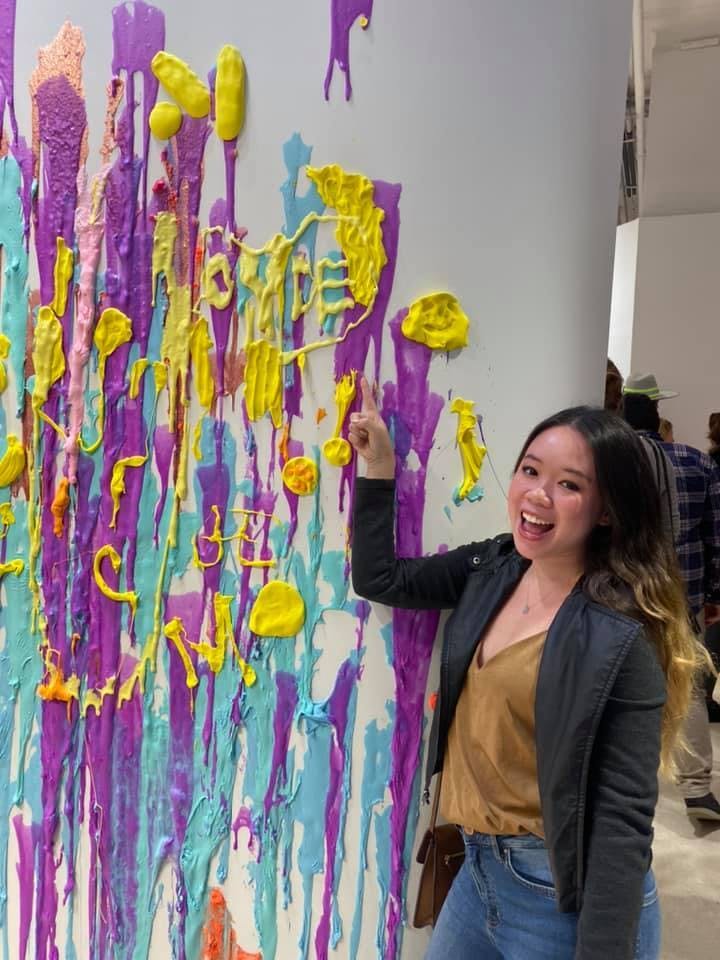 A woman is smiling and pointing at a colorful textured wall art with drips and blobs of yellow, purple, and blue paint.