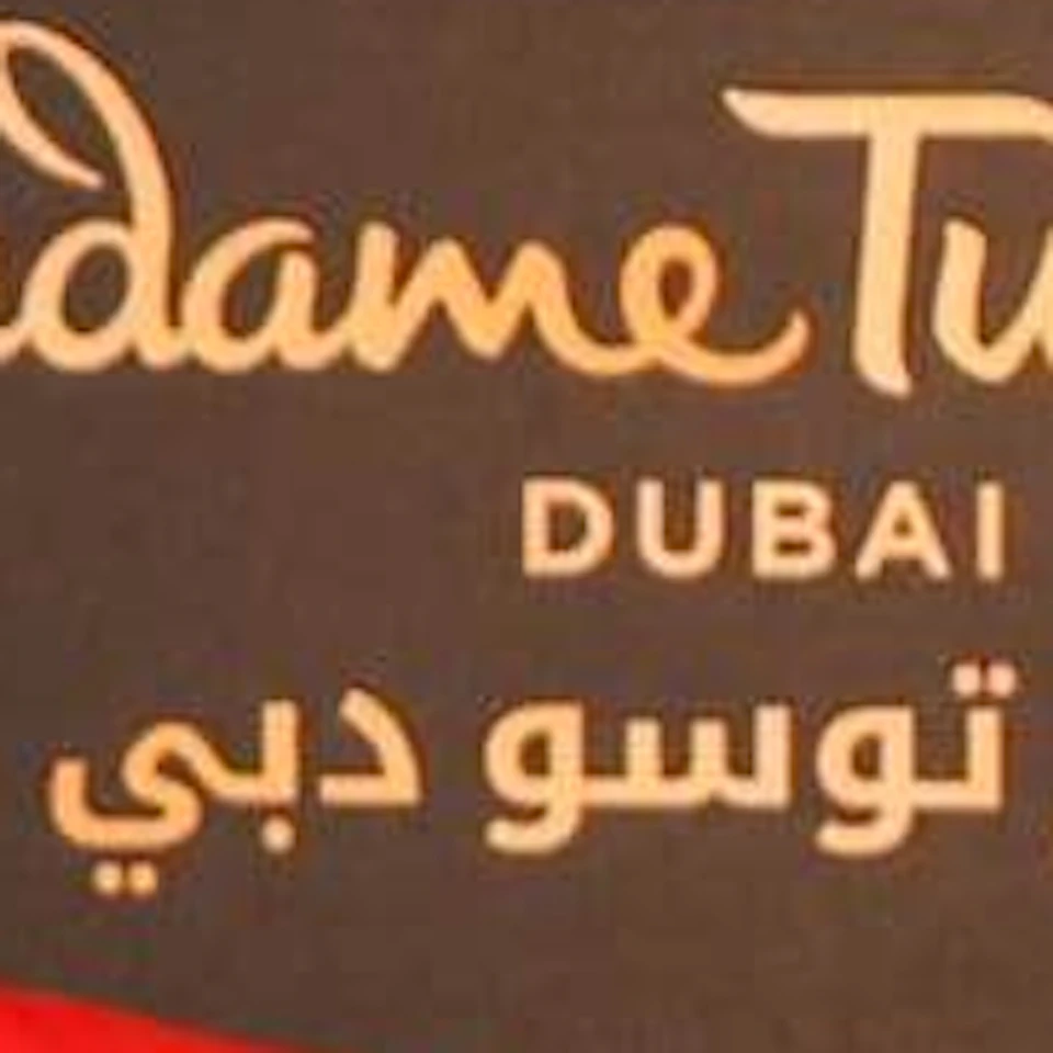 Two men standing in similar poses, both wearing red blazers, white shirts, and black pants, against a Madame Tussauds Dubai backdrop.