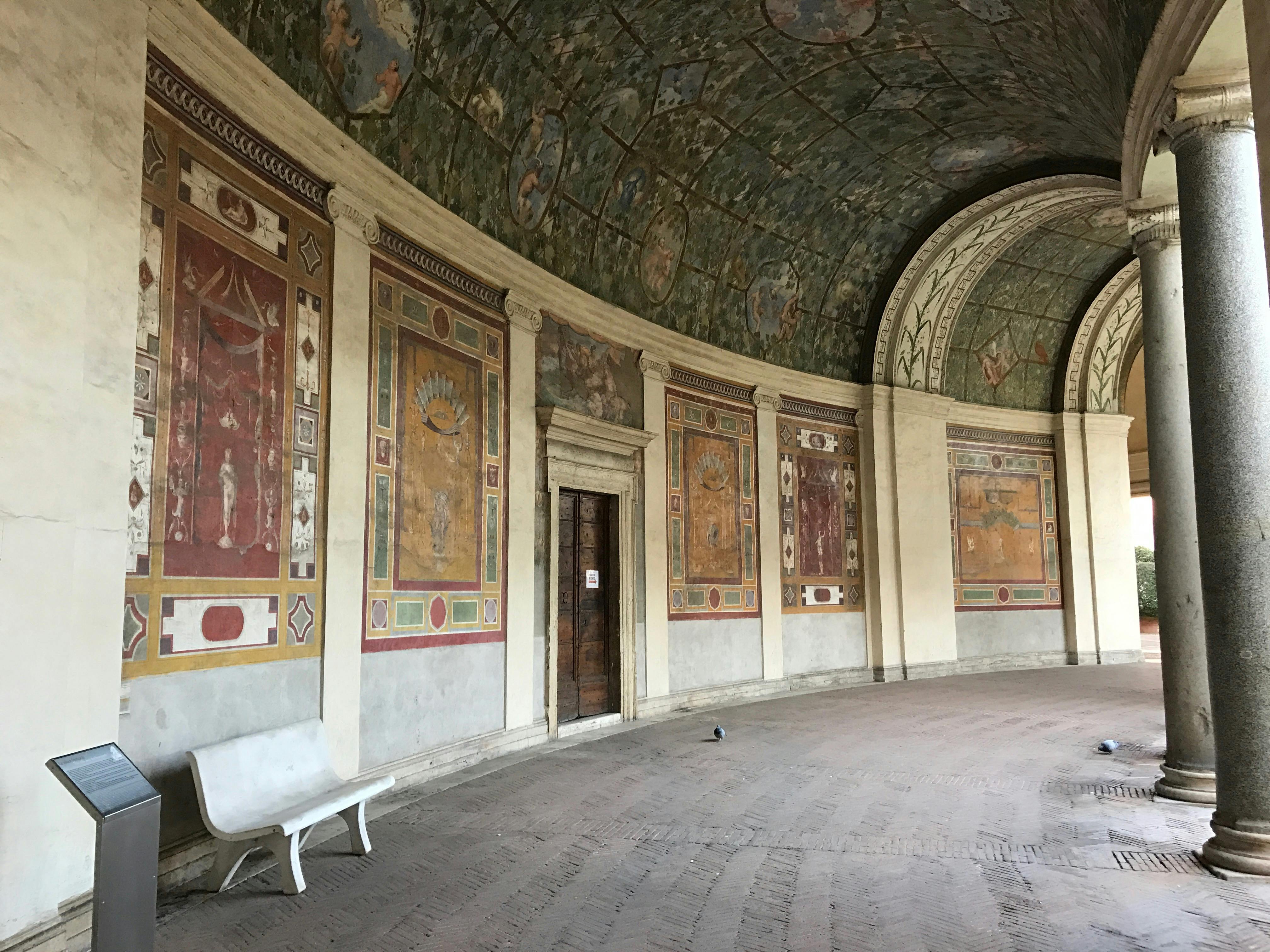 Pièce circulaire avec des peintures murales ornées, un plafond en dôme, une porte en bois et un banc blanc sur un sol carrelé. Un pigeon est posé sur le sol.