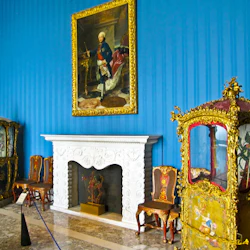 Utsmyckat rum i blått och guld med en vit öppen spis, porträtt, förgyllda stolar och dekorativa vagnar.