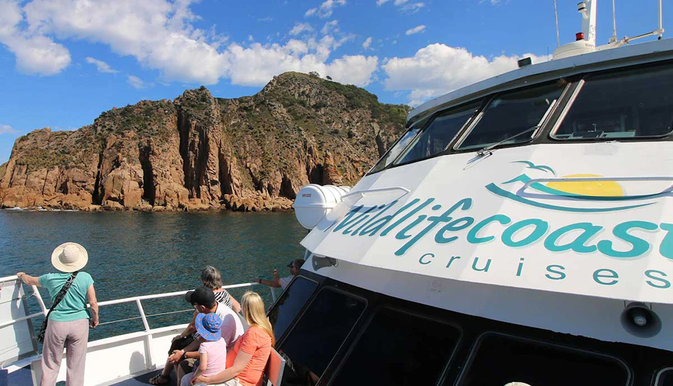 Passengers on a boat tour facing a rugged, rocky coastline under a partly cloudy sky.