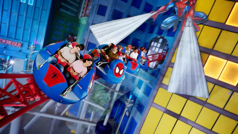People are enjoying a themed amusement park ride featuring Spider-Man, with colorful buildings and tracks visible in the background.