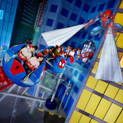People are enjoying a themed amusement park ride featuring Spider-Man, with colorful buildings and tracks visible in the background.