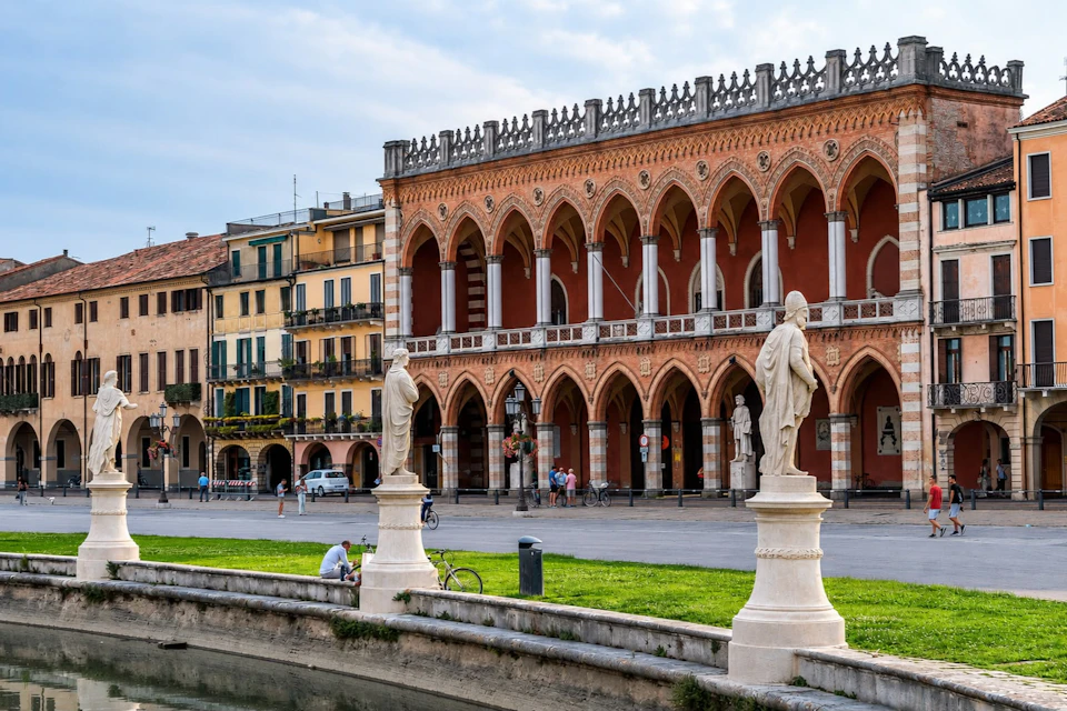 A historic building with arches and statues in front, surrounded by grass and a canal in a city setting.