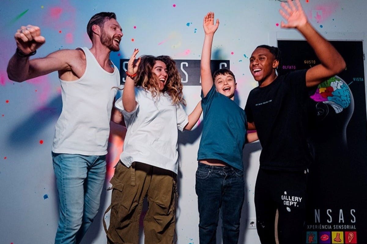 Quattro persone sorridono e alzano le mani in segno di festa su una parete colorata con testo e grafica.