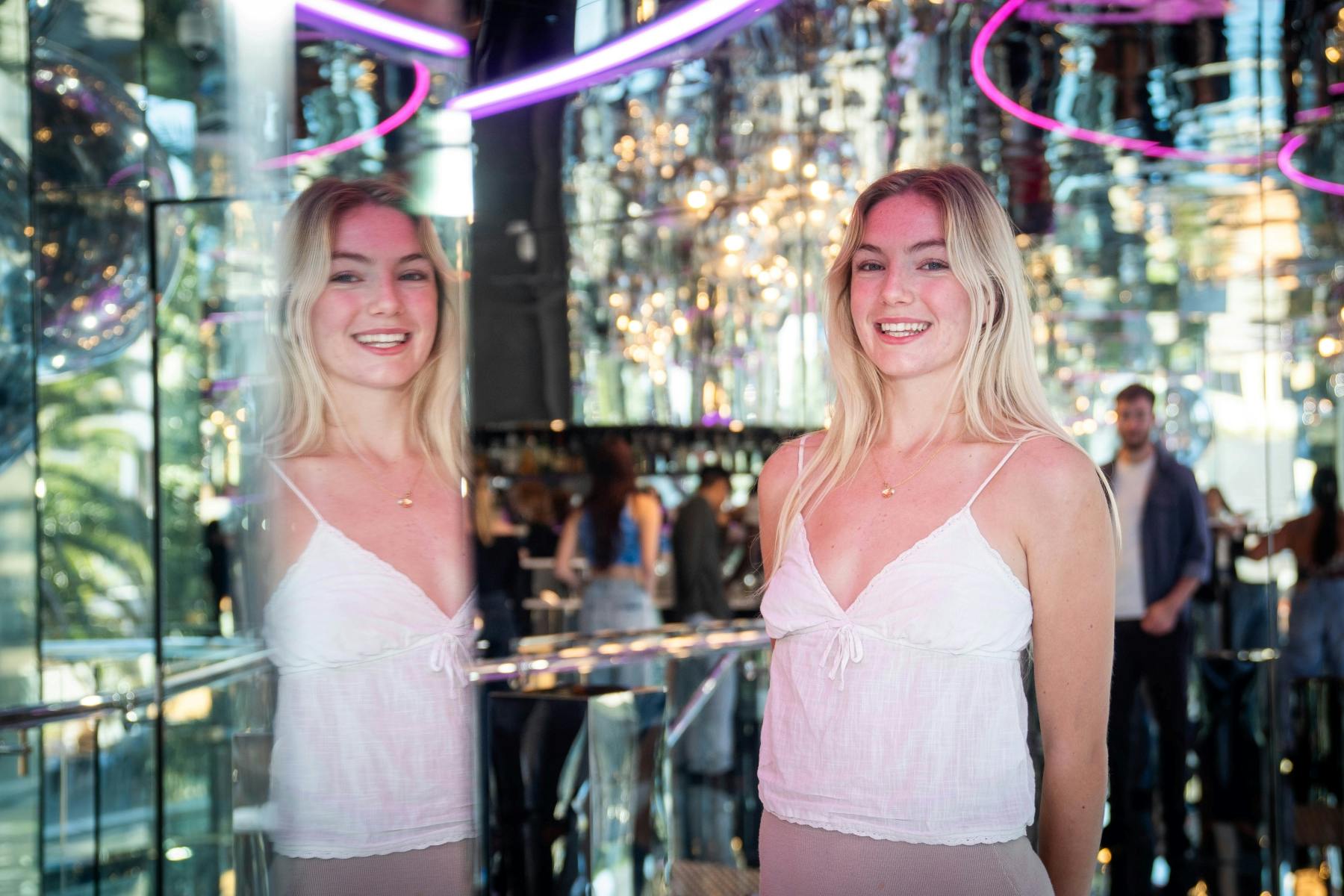 白い服を着た女性が、反射面、カラフルな光、背景にぼやけた人々がいる部屋で微笑んでいます。