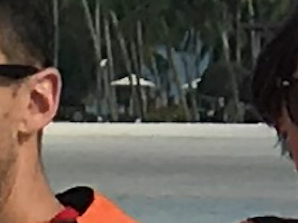 Two people ride a jet ski on the ocean, one with an arm raised. Another jet ski is seen behind. Mountains and trees are in the background.
