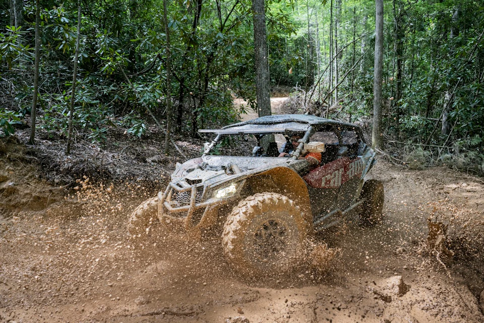 An off-road vehicle driving through a muddy forest trail, splashing mud, surrounded by dense green trees.
