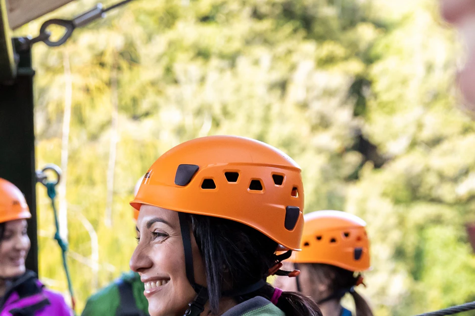 People wearing helmets and harnesses prepare for a zip-lining activity on a wooden platform surrounded by forest.