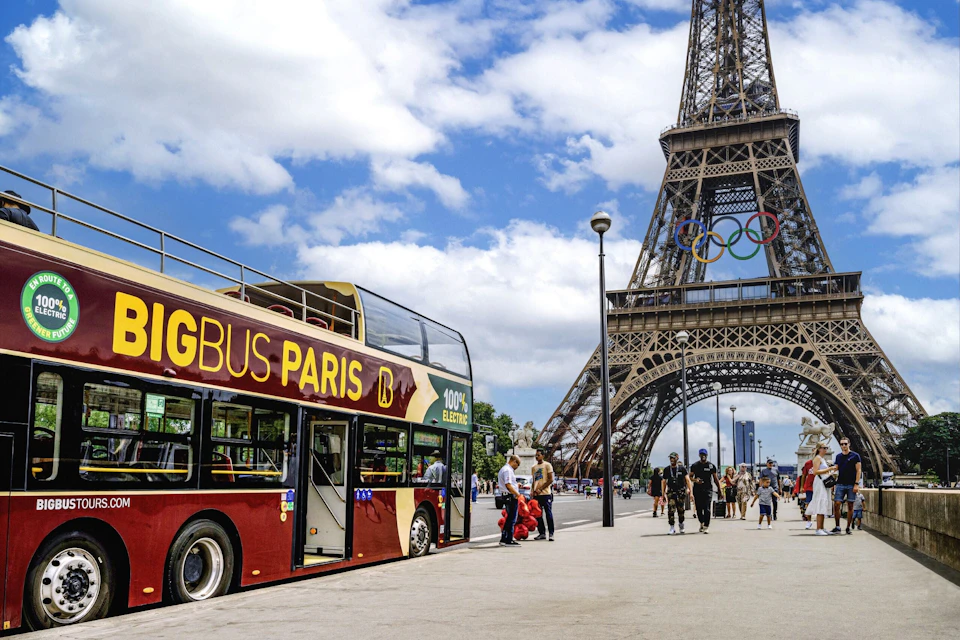 A red double-decker sightseeing bus near the Eiffel Tower adorned with Olympic rings under a partly cloudy sky.