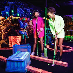 Två kvinnor spelar neonminigolf inomhus, skrattar och håller klubbor. Bakgrunden har en levande, glödande inredning med pirattema.