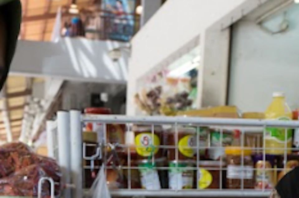 Two women interact at a market stall filled with various spices and packaged goods. One woman holds a small packet.
