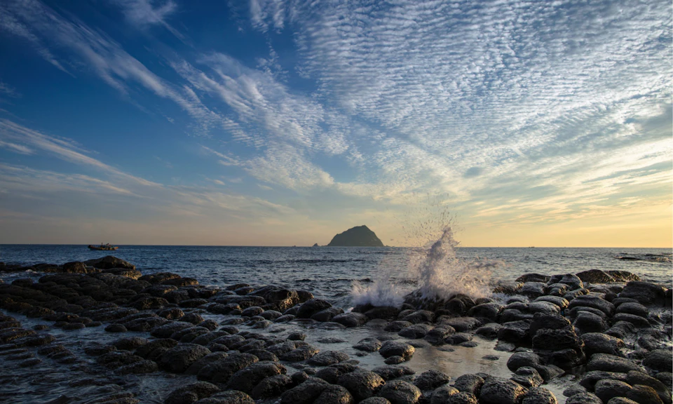 Waves splashing onto a rocky shore under a cloudy sky, with an island visible in the distance during sunset.