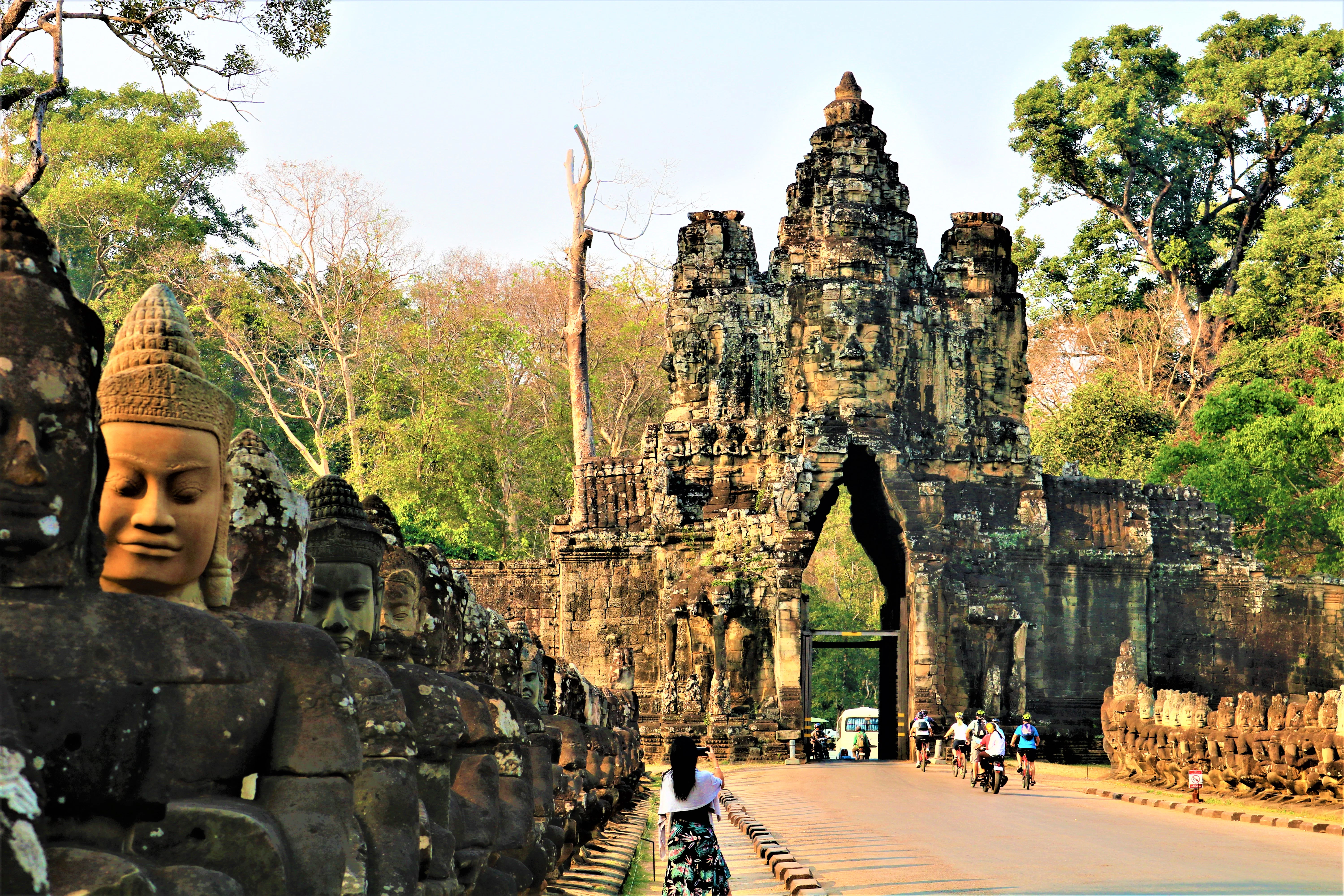 A line of stone Buddha statues leads to an ancient stone gateway; a woman in white and black stands near the entrance.