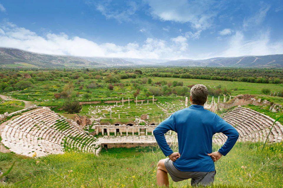 A person in a blue shirt overlooks ancient ruins with columns and stone structures, surrounded by green fields and a cloudy sky.