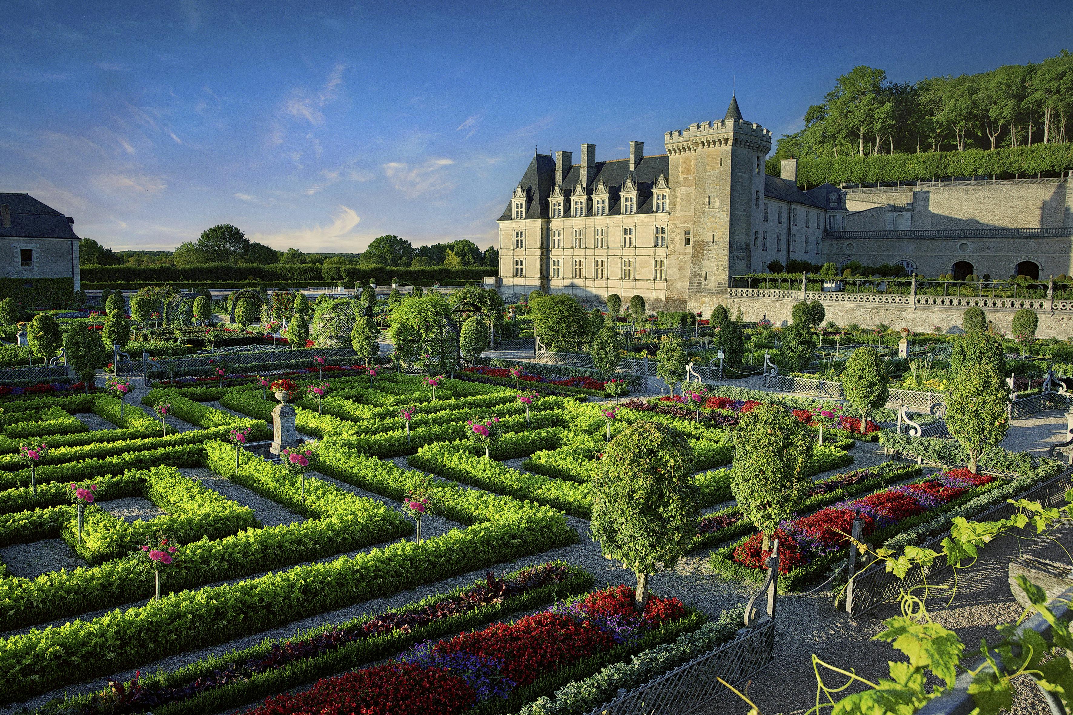 Giardini ornati con siepi curate e fiori colorati di fronte a un grande castello storico sotto un cielo azzurro e limpido.