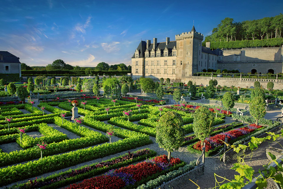 Manicured gardens with colorful flowerbeds and hedges in front of a historic stone castle under a clear blue sky.