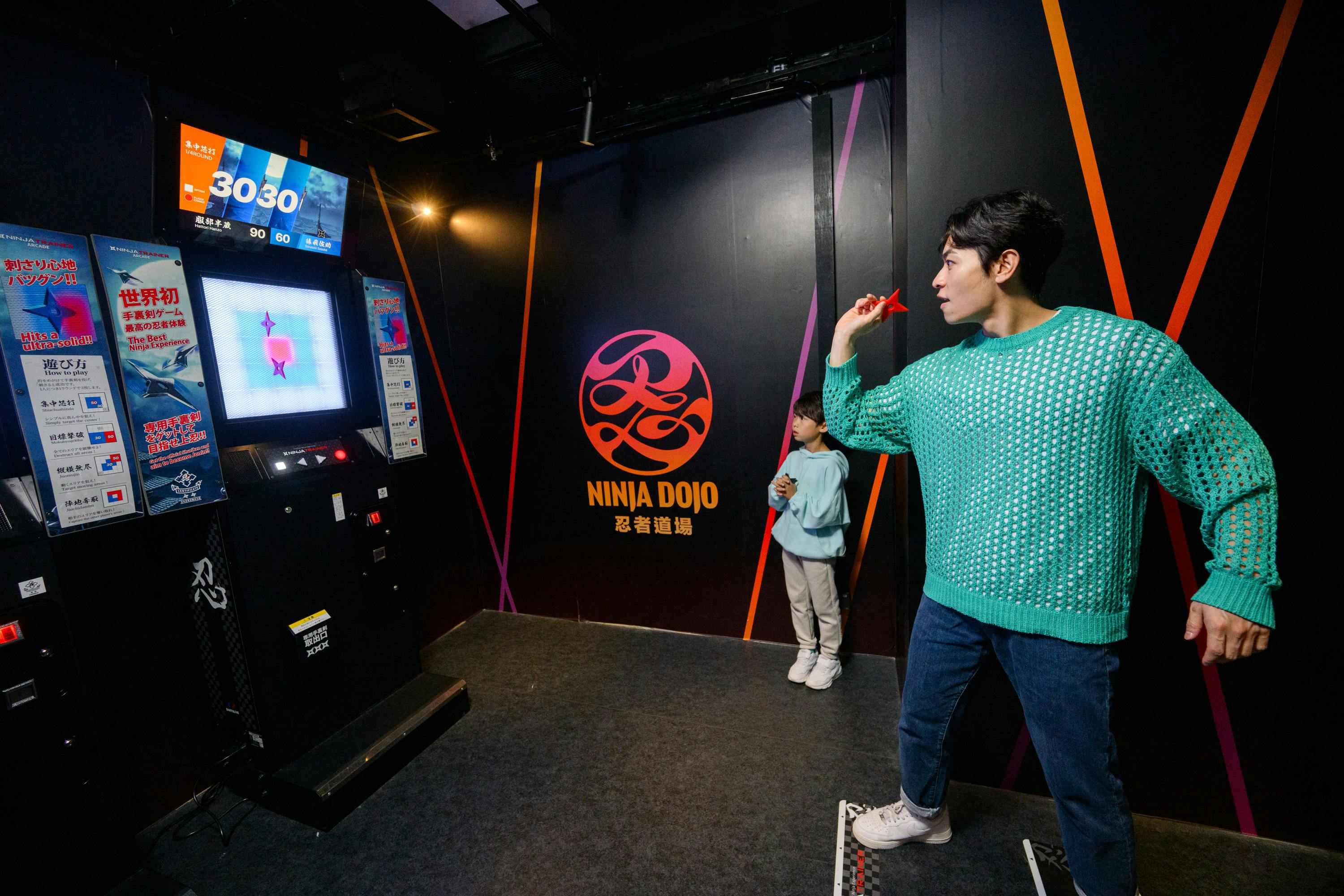 A man and a child stand in a room with a "Ninja Dojo" logo on the wall. The man is holding a red throwing star.