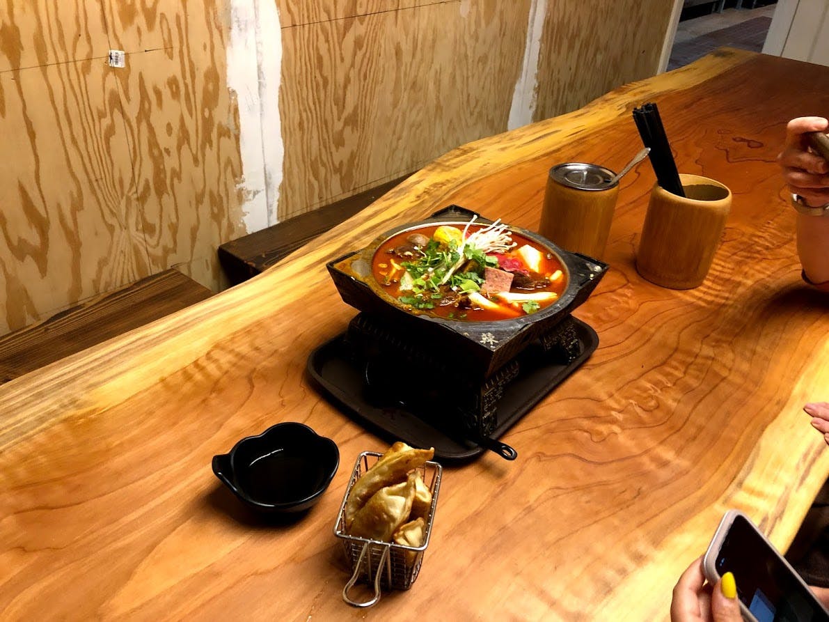 Een pot hotpotsoep met verschillende ingrediënten op een houten toonbank, vergezeld van een klein mandje gefrituurde knoedels en dipsaus.