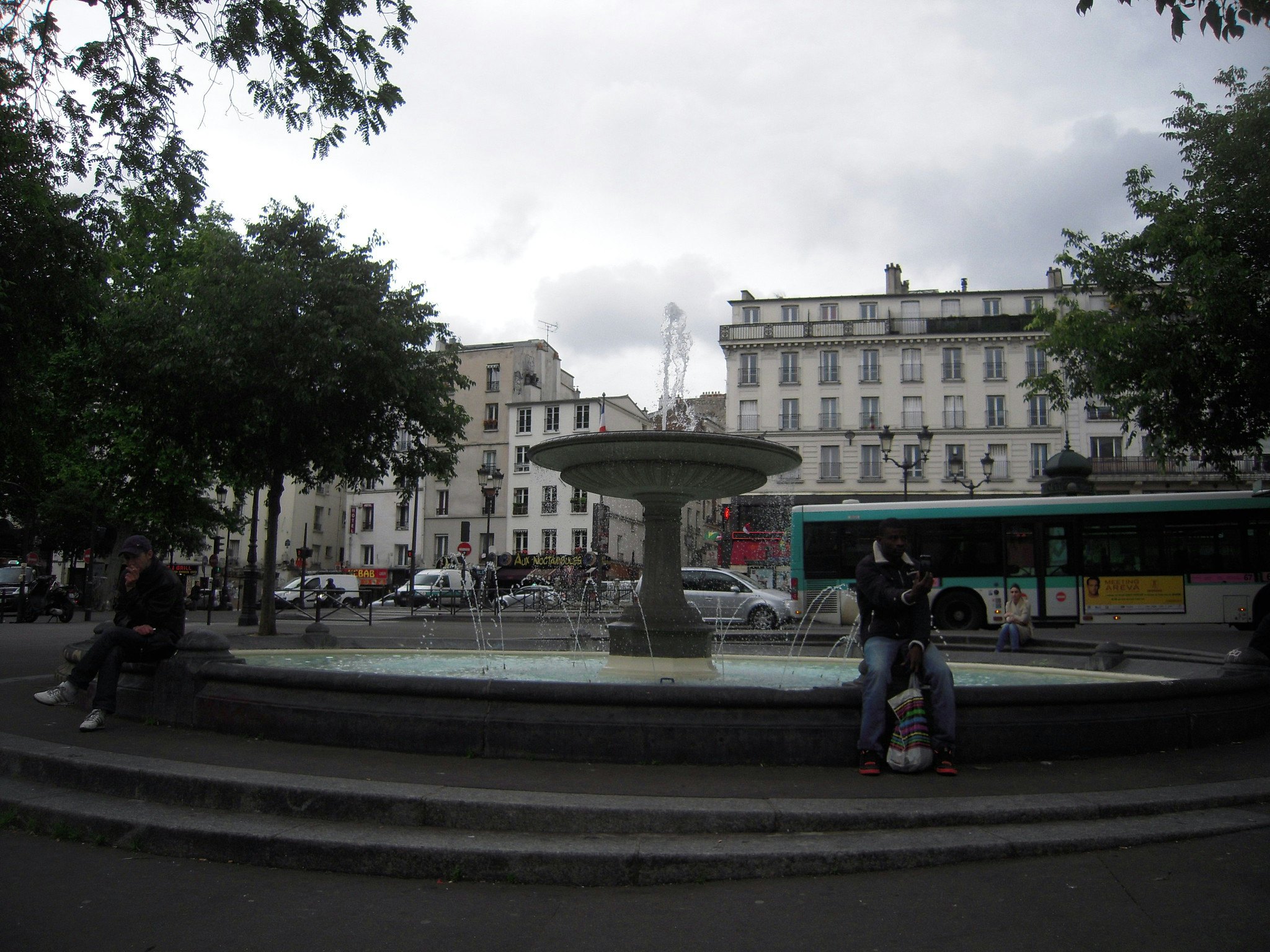 Pessoas sentadas à volta de uma fonte numa praça urbana, com edifícios, árvores e um autocarro como pano de fundo num dia nublado.