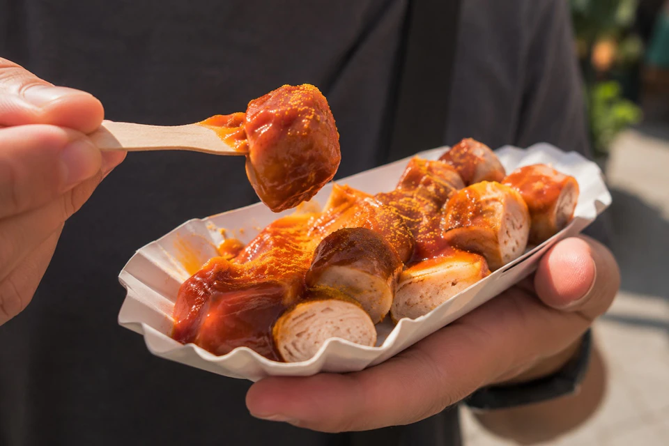 A person holding a tray of sliced sausage topped with curry ketchup, using a wooden fork to pick up a piece.