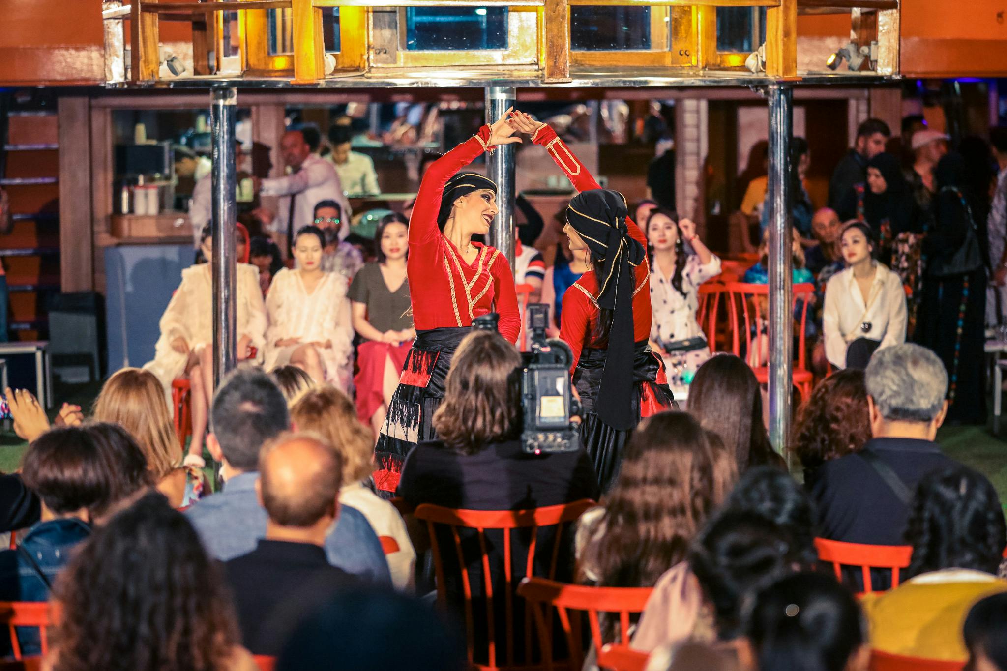 赤いコスチュームを着た2人のダンサーが、着席した観客の前でカラフルな室内環境で演奏します。