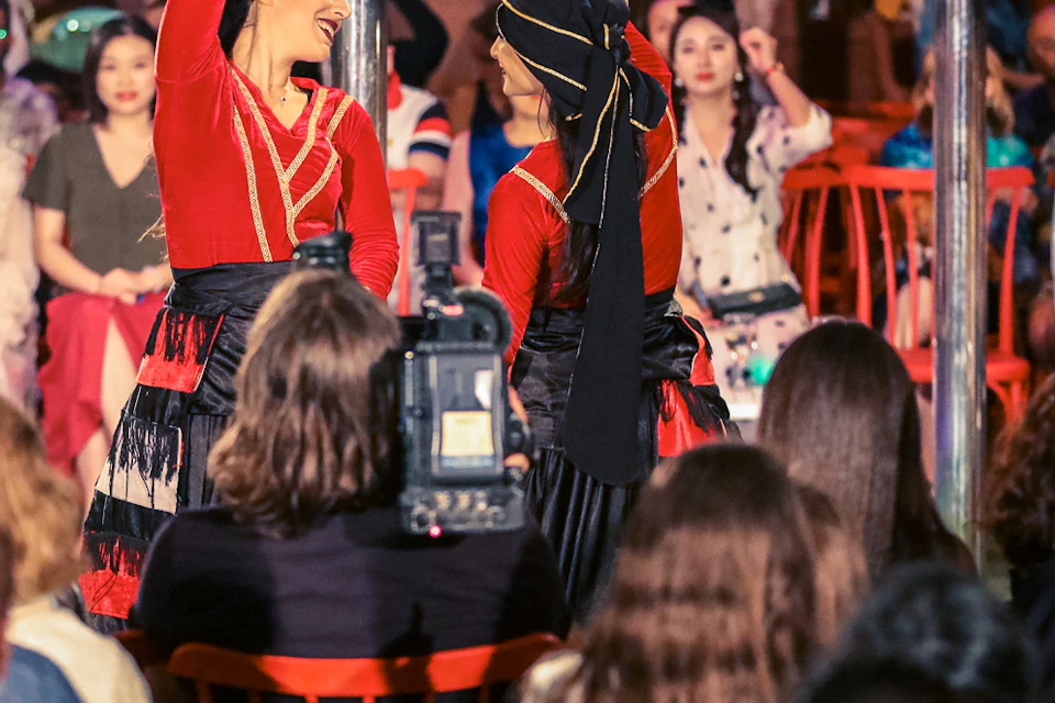 Two dancers in red costumes perform in front of a seated audience in a colorful indoor setting.