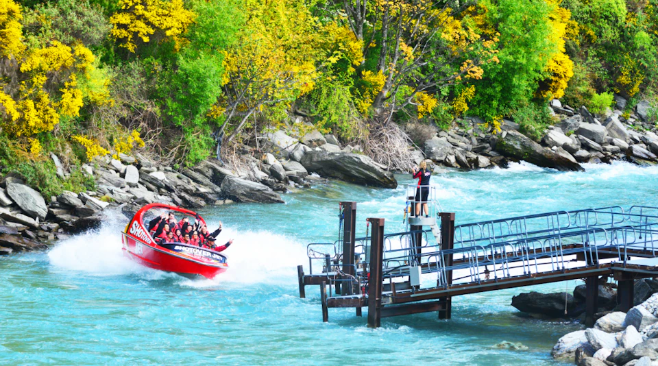 A red jet boat speeding on a blue river near a dock, surrounded by trees with yellow and green foliage.