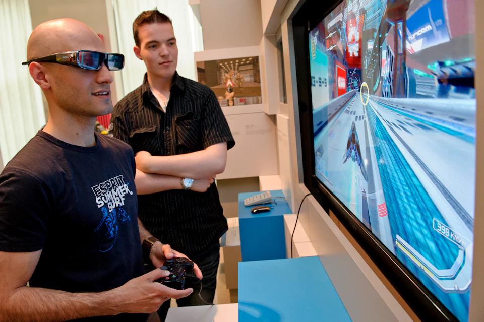 Two people are playing a video game. One wears glasses and holds a controller, while the other watches with arms crossed.