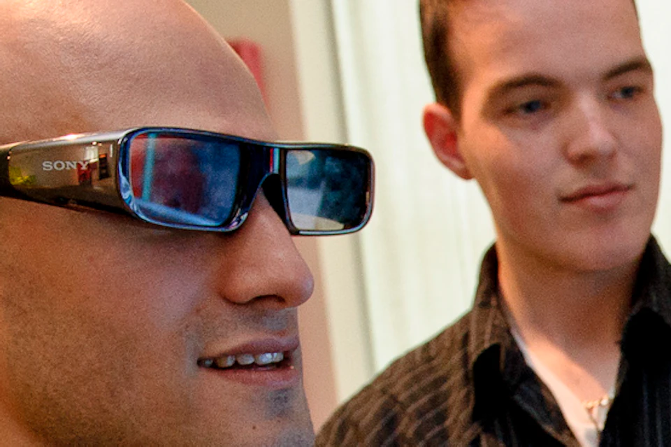 Two people are playing a video game. One wears glasses and holds a controller, while the other watches with arms crossed.