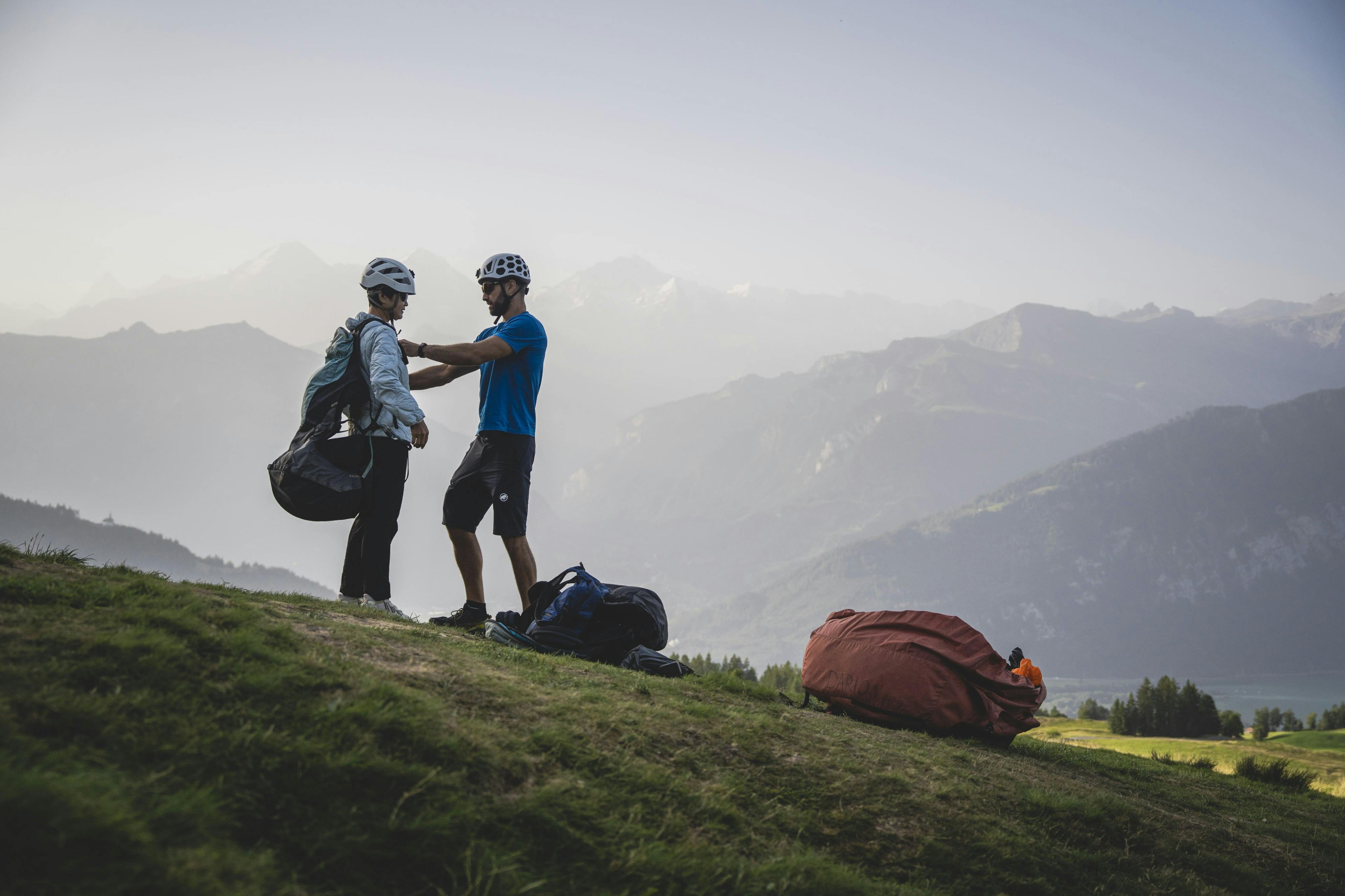Deux personnes casquées préparent du matériel sur une colline herbeuse, avec un paysage montagneux en arrière-plan.